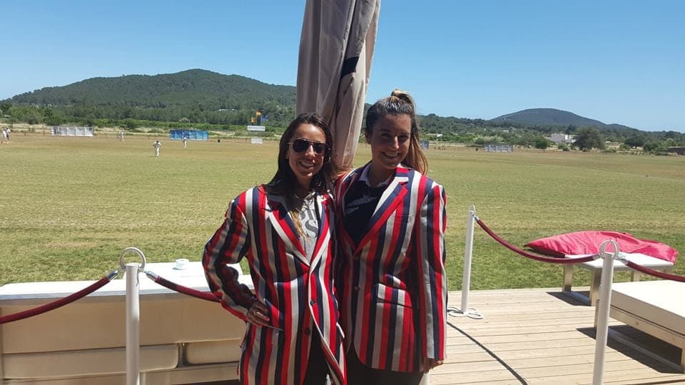 Two LNZCC members in striped club blazers beside a sunny cricket ground with hills behind